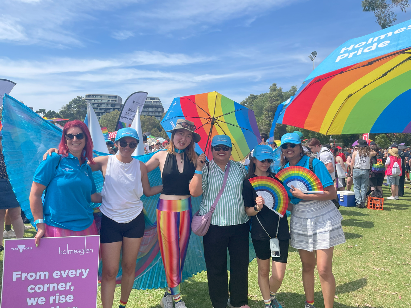 Six smiling staff stand together holding rainbow fans and a sign reading 