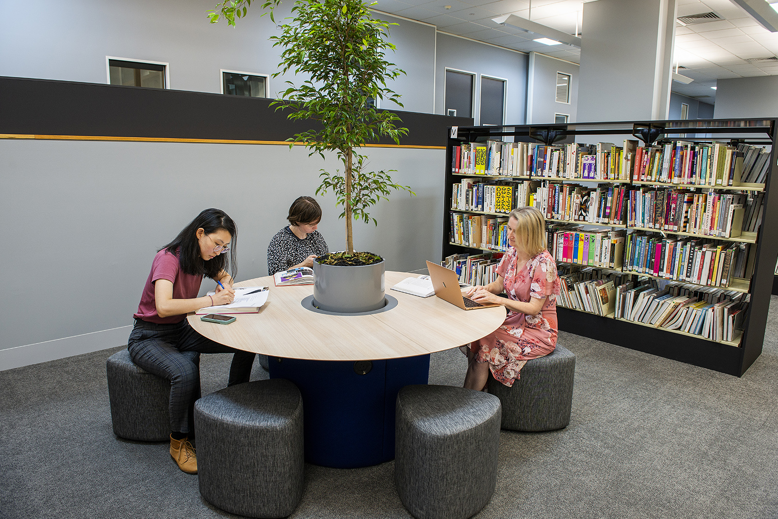 Three people sitting around a table in front of a bookshelf using books and a laptop
