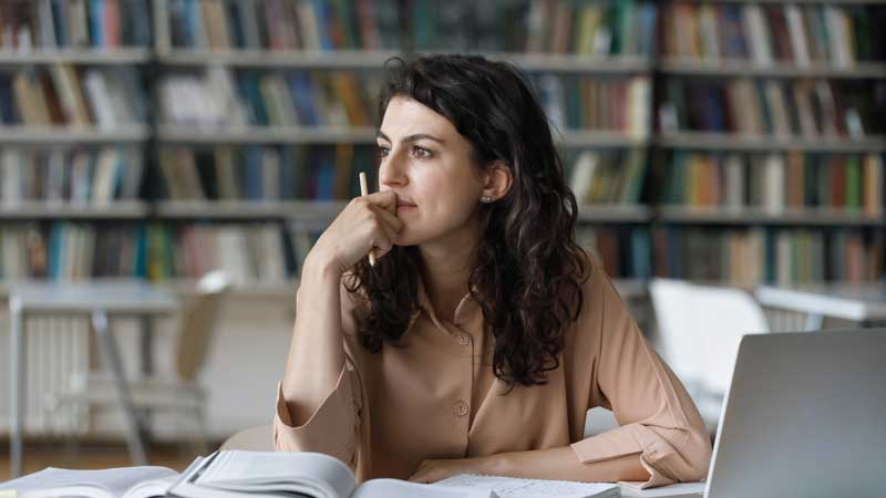 A person sitting at a table in a library, looking thoughtfully off to the side